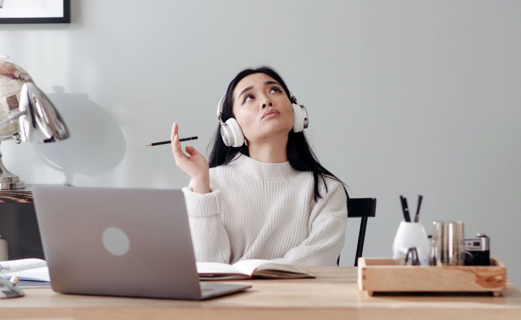 A woman sitting at her desk, wearing headphones. She is holding a pen and there are various office items on her desk. Her laptop is open in front of her. She is considering the features she would like her cannabis marketing software to have for her dispensary.  